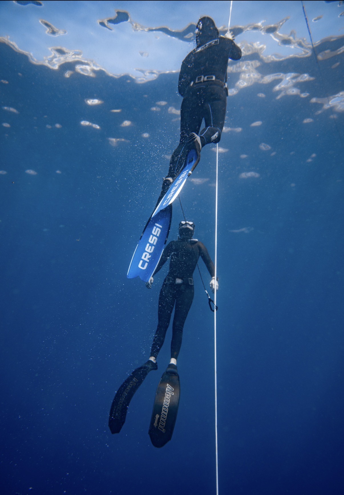 Two freedivers ascending the line together in deep blue water, one with blue Cressi fins