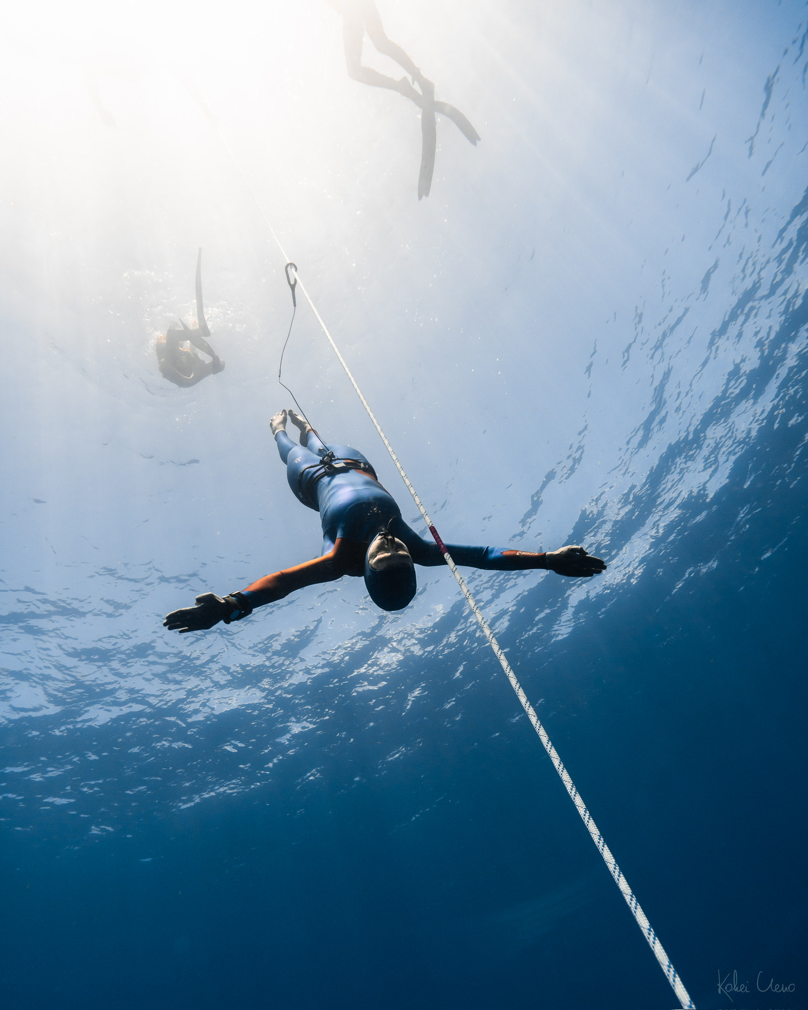 Natalie Bruce hanging at the freediving line beneath a sunburst, a safety diver above in clear blue water